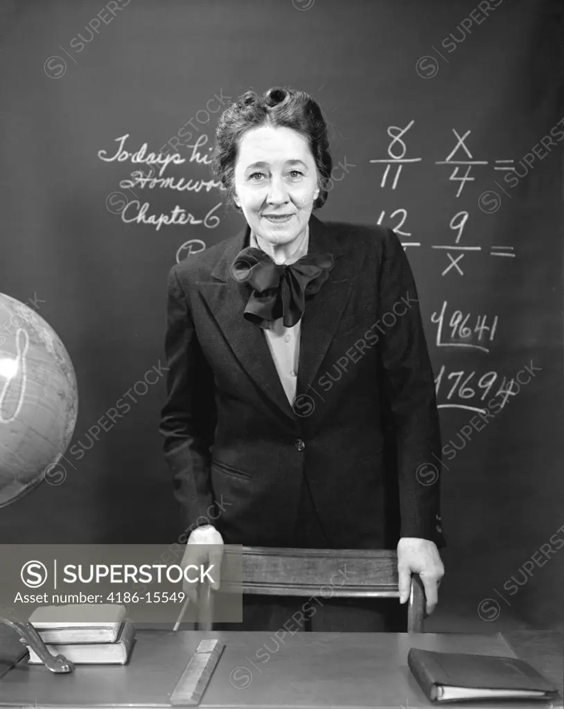 1940S Smiling School Teacher Standing Behind Her Desk & In Front Of The Black Board With Fractions