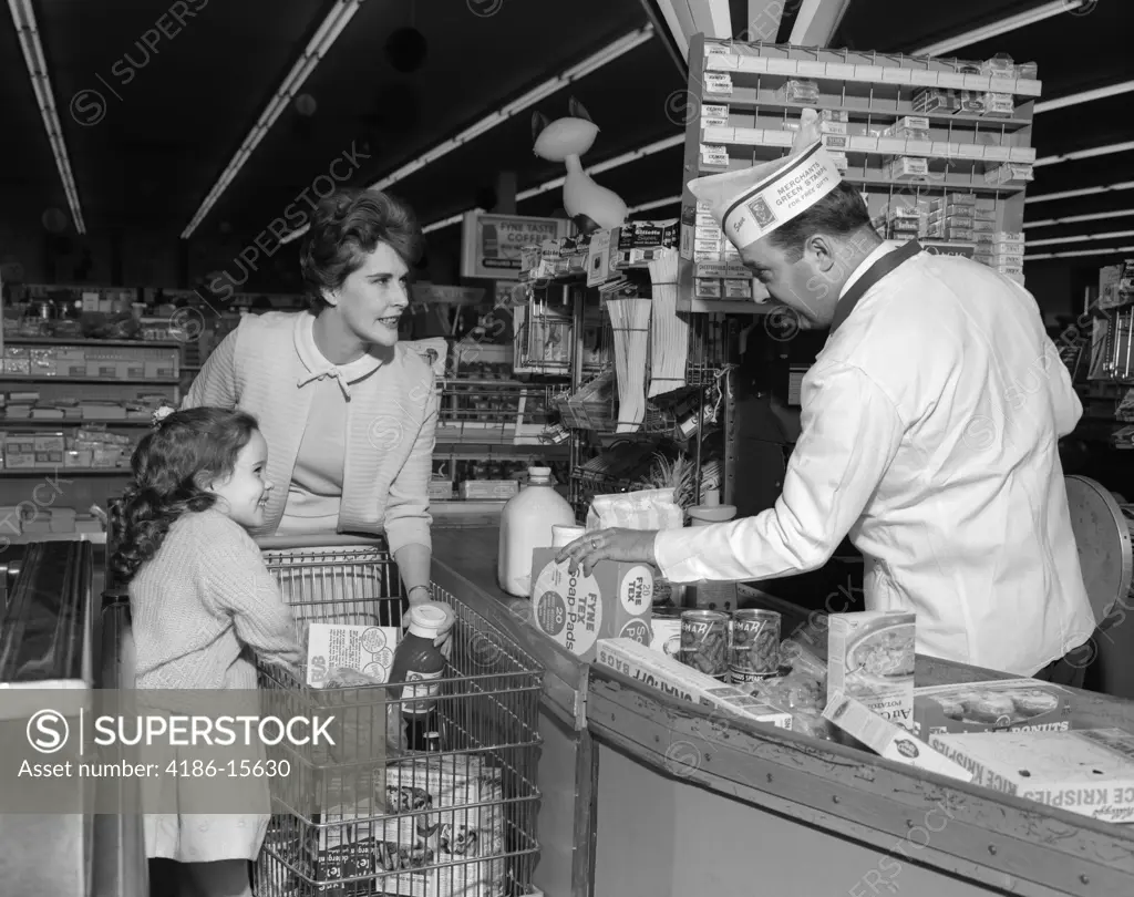 1960S Mother Daughter Unload Grocery Cart At Supermarket Checkout Counter Man Clerk Cashier Rings Up Cash Register