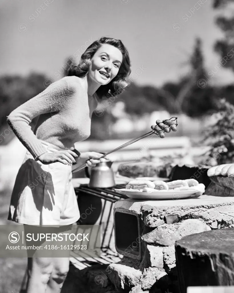 1950s SMILING BRUNETTE WOMAN WEARING SHORT SHORTS SWEATER COOKING HOT DOGS  OVER OUTDOOR STONE GRILL LOOKING AT CAMERA - SuperStock