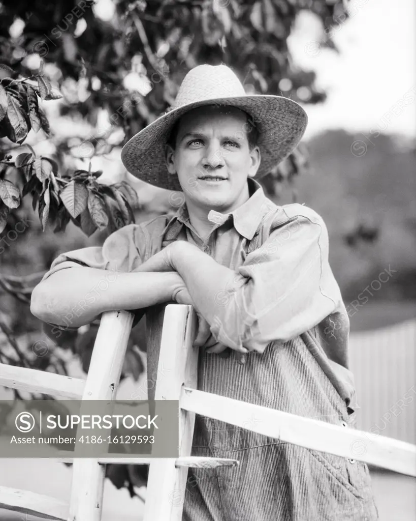 1920s-1930s Farm Boy Wearing Straw Hat And Overalls Sitting On Log