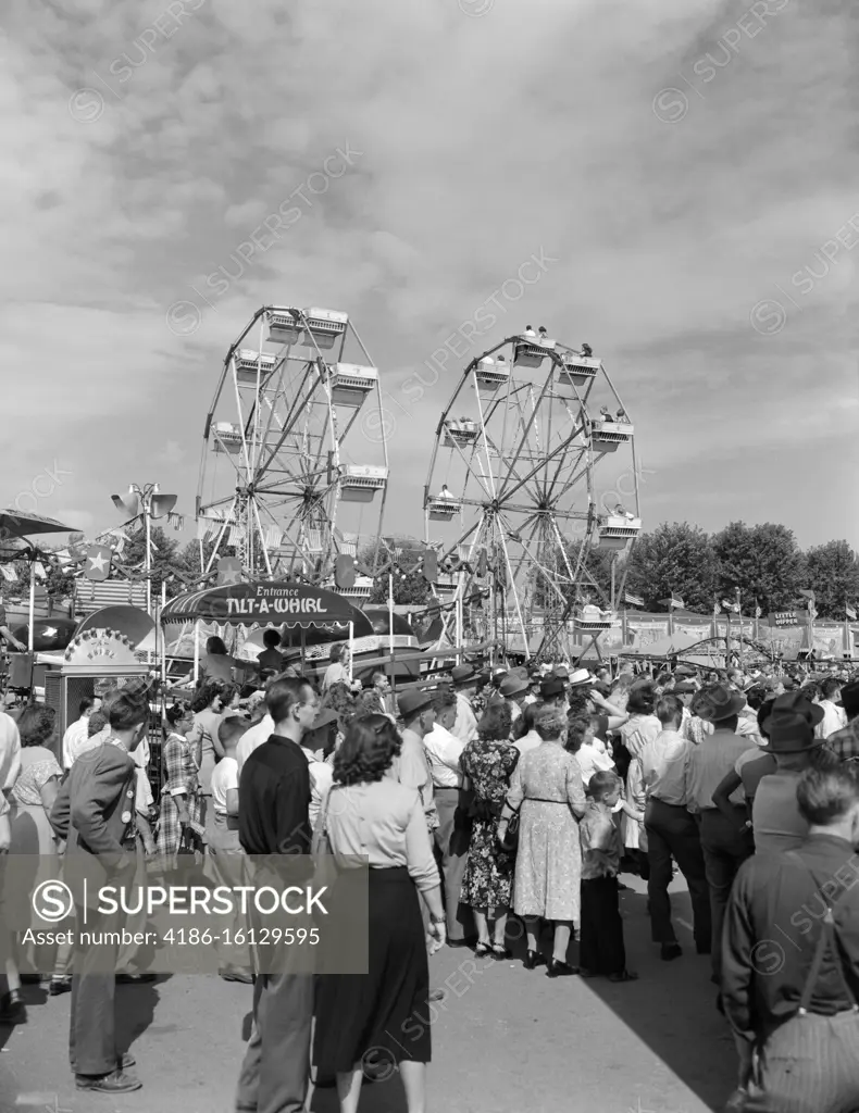 1940s 1950s CROWD AT LOCAL COUNTY FAIR CARNIVAL WITH 2 FERRIS WHEELS IN BACKGROUND