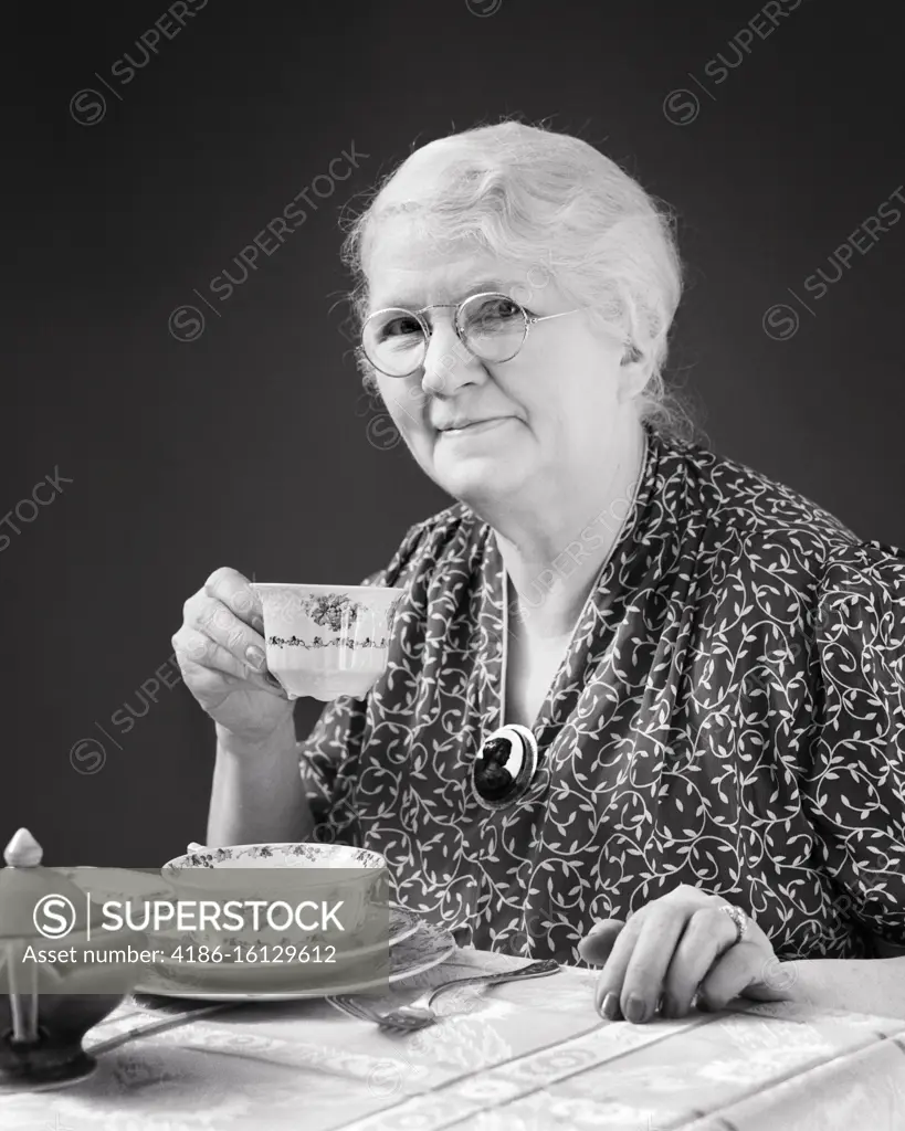 1940s SENIOR WOMAN WEARING EYEGLASSES CAMEO BROOCH DRINKING CUP COFFEE TEA SITTING AT DINING TABLE LOOKING AT CAMERA