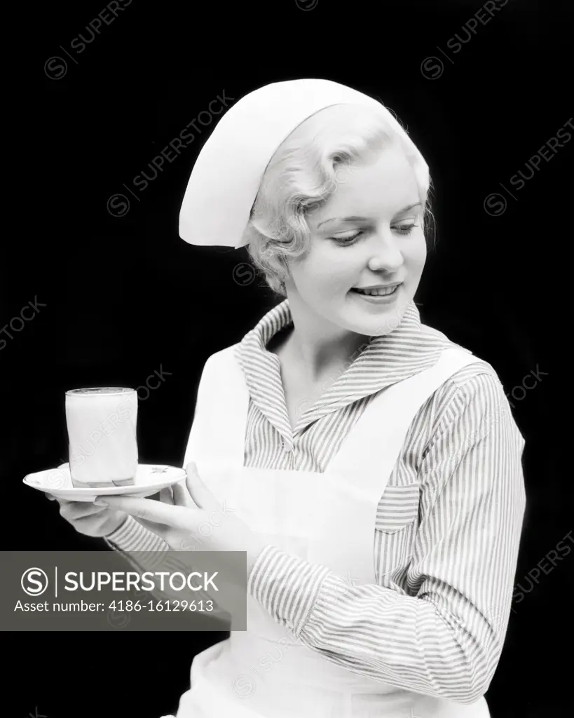 1930s PRETTY BLONDE NURSE HOLDING A DISH WITH GLASS OF MILK LOOKING DOWN TO TALK TO A PATIENT