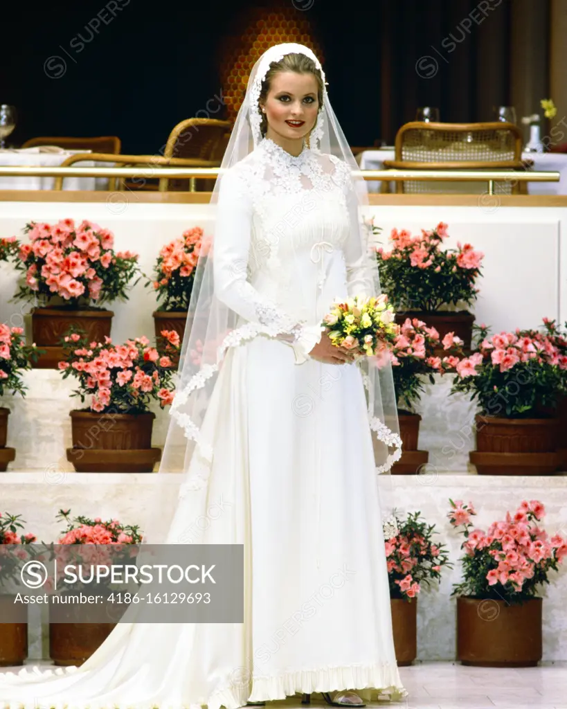 1970s 1980s SMILING BRIDE IN GOWN LOOKING AT CAMERA HOLDING YELLOW BOUQUET LONG VEIL LACE TRIM STANDING BY TIERS OF PINK AZALEAS