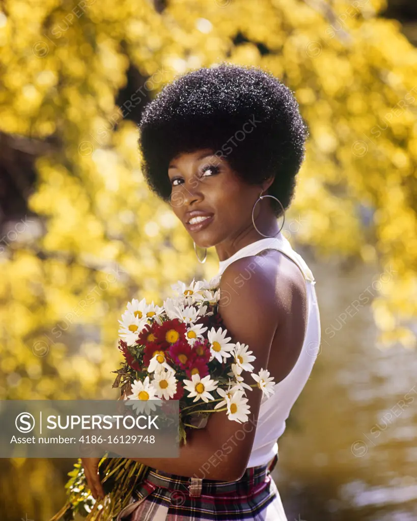 1970s SMILING AFRICAN AMERICAN WOMAN WEARING HOOP EARRINGS WHITE TANK TOP HOLDING A BOUQUET SPRING FLOWERS LOOKING AT CAMERA