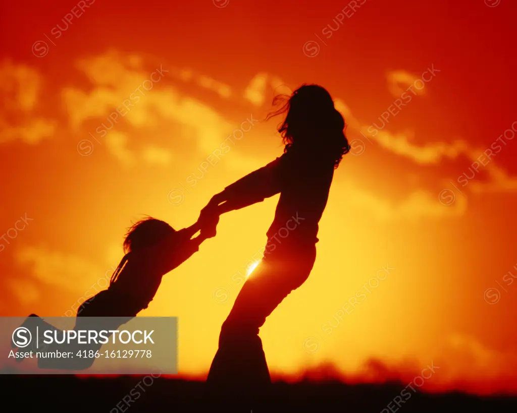 1970s 1980s AN ANONYMOUS SILHOUETTED JOYOUS MOTHER SWINGING CHILD GIRL OR BOY BY BOTH ARMS IN GOLDEN LIGHT SUNRISE OR SUNSET 