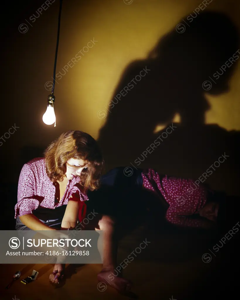 1960s 1970s FEMALE DRUG ADDICT SITTING UNDER BARE BULB MAINLINING INJECTING DRUGS HEROIN MALE COMPANION SLEEPING ON THE FLOOR 