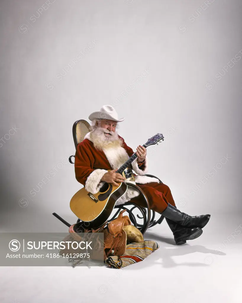 1980s COWBOY SANTA CLAUS LOOKING AT CAMERA SITTING IN BENTWOOD ROCKER BESIDE WESTERN SADDLE PLAYING GUITAR WEARING WHITE HAT