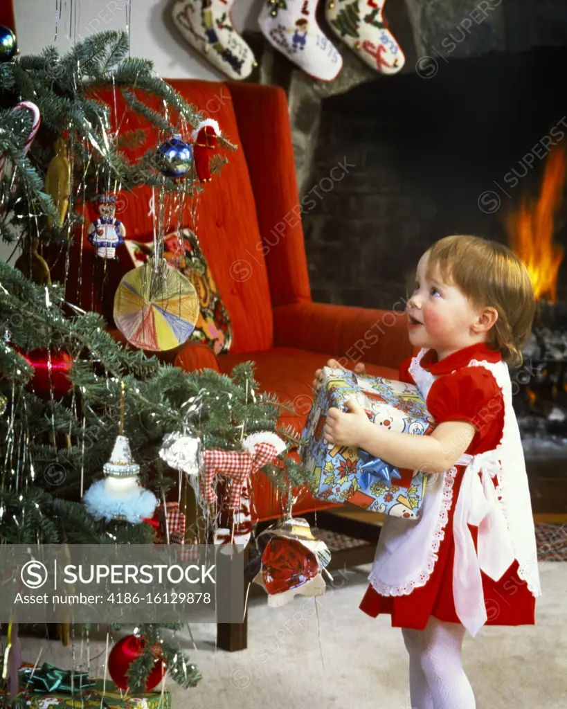 1970s AWESTRUCK LITTLE GIRL TODDLER HOLDING GIFT WRAPPED PRESENT LOOKING UP AT DECORATED CHRISTMAS TREE BY FIREPLACE