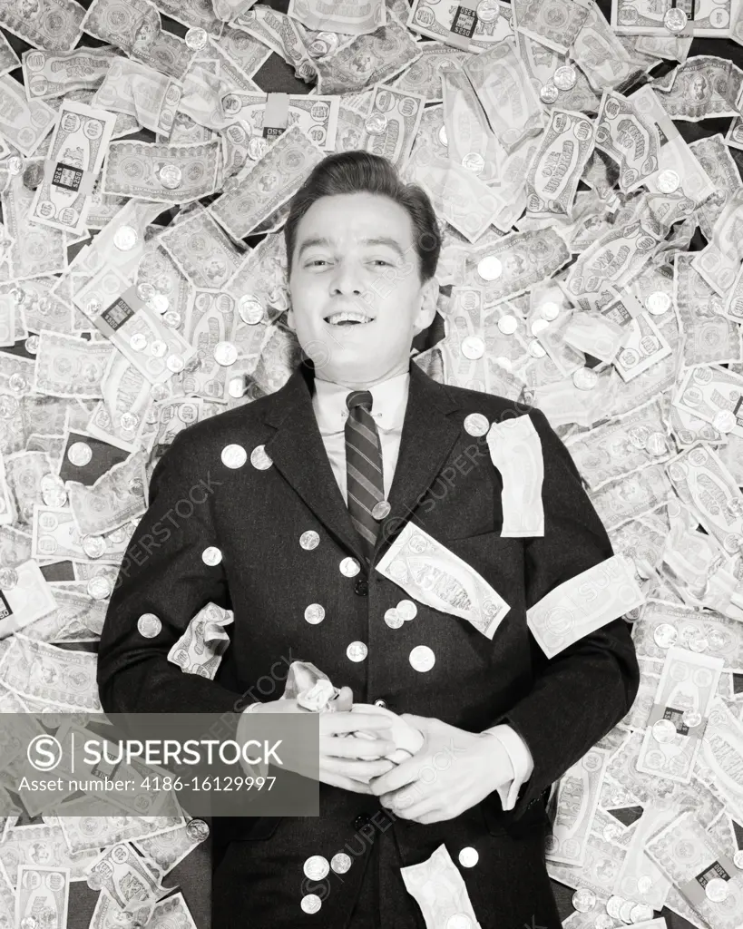 1950s SMILING  MAN LYING ON BACK ON A PILE OF MONEY COINS AND BILLS LOOKING AT CAMERA WINNER LUCKY LOTTERY PRIZE MONEY