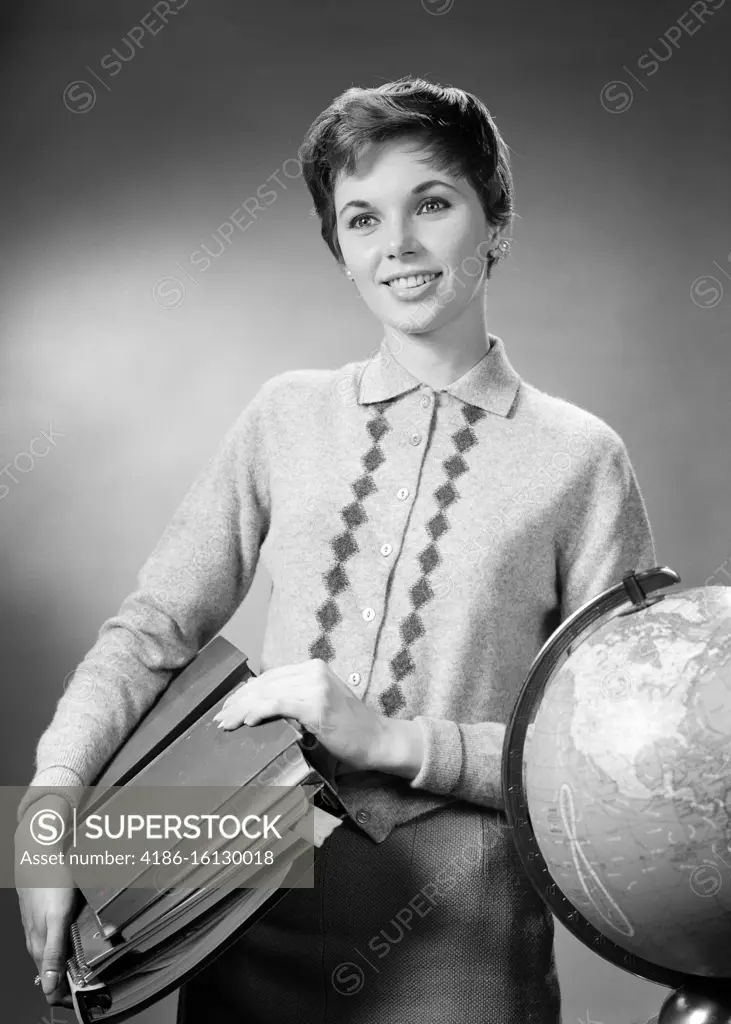 1950s 1960s  BRUNETTE WOMAN TEACHER CARRYING CLASS MATERIALS  BOOKS NOTEBOOKS SMILING STANDING BY GLOBE WEARING SWEATER