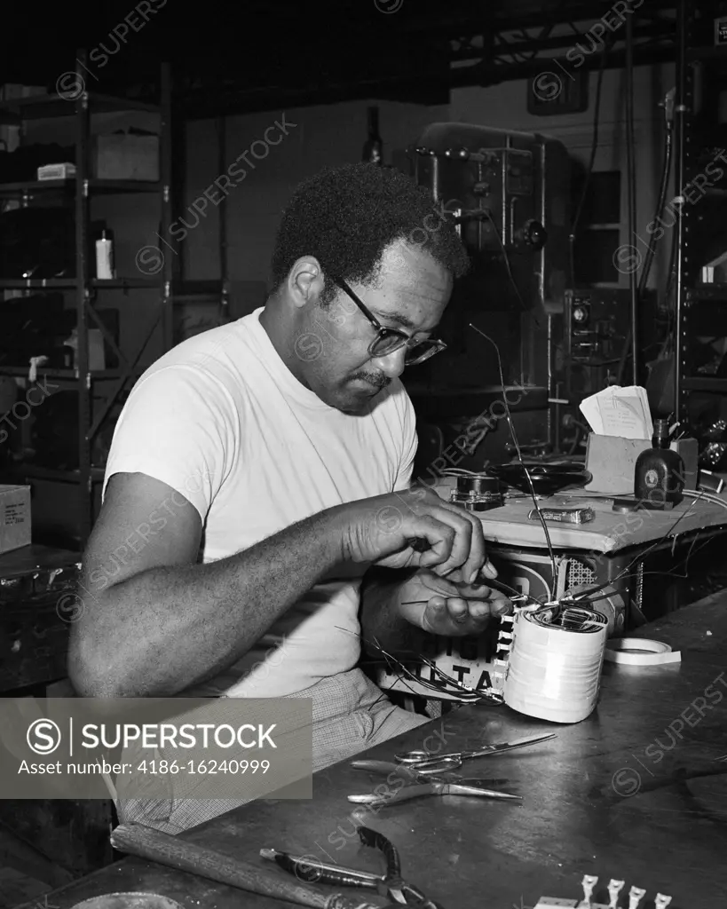 1960s 1970s SKILLED MAN AFRICAN-AMERICAN ELECTRICAL TECHNICIAN REBUILDING REPAIRING AN INDUSTRIAL MACHINE COMPONENT IN SHOP