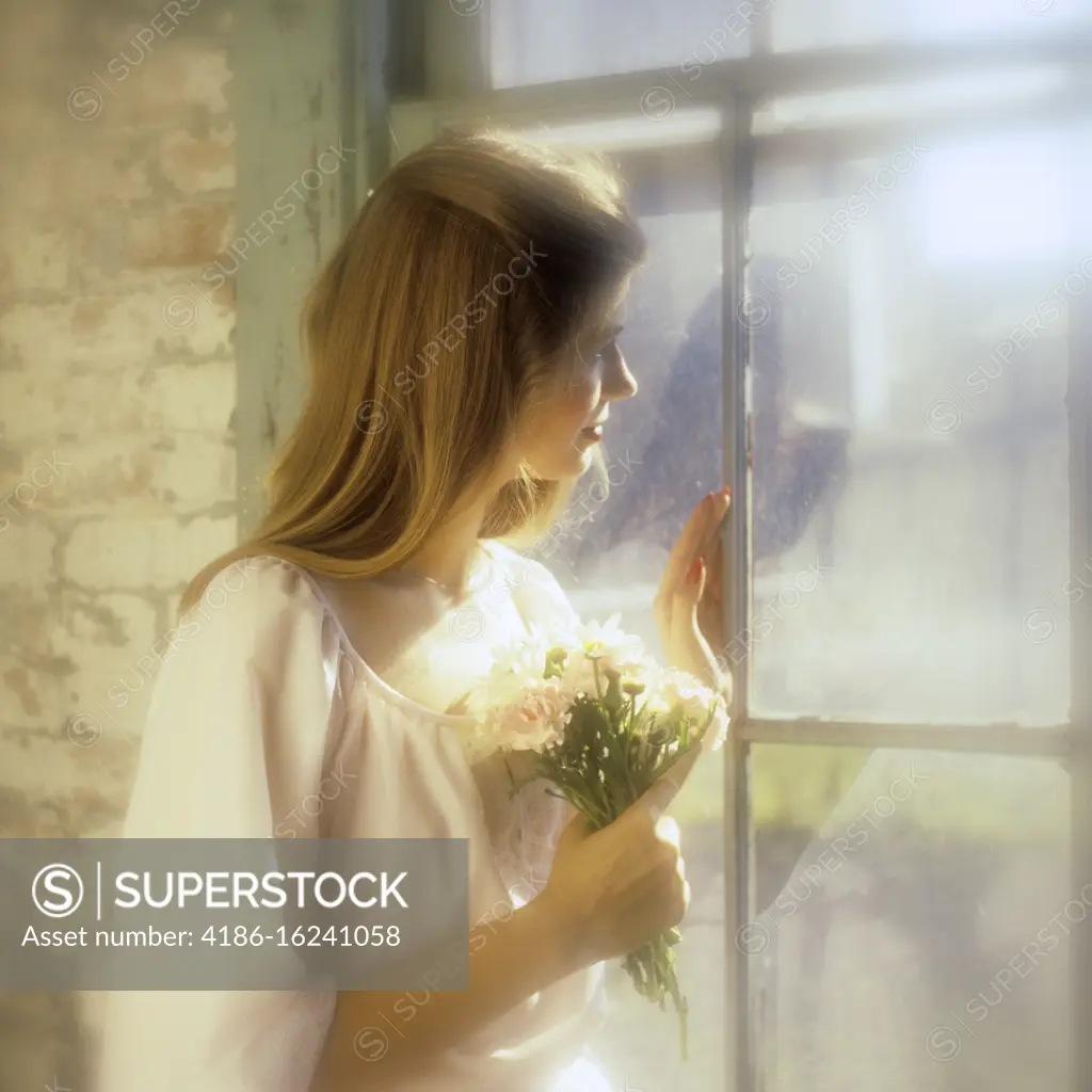 1970s PENSIVE WOMAN WEARING COTTON PEASANT DRESS HOLDING BOUQUET OF WILD FLOWERS LOOKING OUT OF SUNNY WINDOW IN OLD BRICK HOUSE 