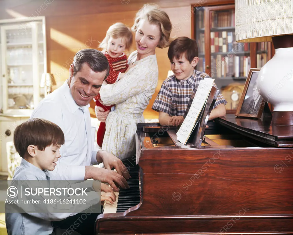 1960s 1970s FATHER AND SON PLAYING PIANO WITH MOTHER HOLDING DAUGHTER AND BROTHER WATCHING FAMILY MUSIC IN LIVNG ROOM