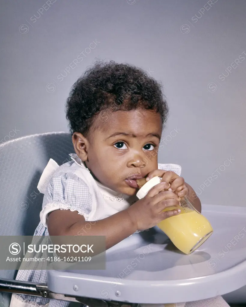 1960s WIDE-EYED AFRICAN AMERICAN BABY GIRL LOOKING AT CAMERA SITTING IN HIGH CHAIR DRINKING ORANGE JUICE FROM GLASS BOTTLE