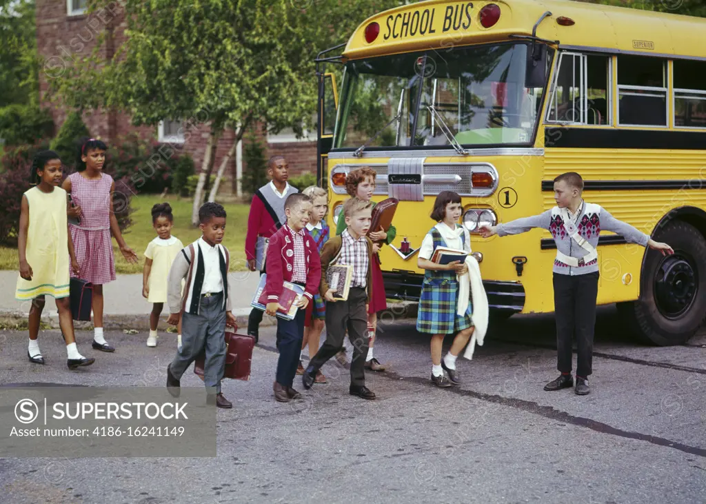 1960s ETHNICALLY MIXED GROUP OF SCHOOL CHILDREN BOYS GIRLS CROSSING STREET BY SCHOOL BUS ASSISTED BY SAFETY PATROL BOY