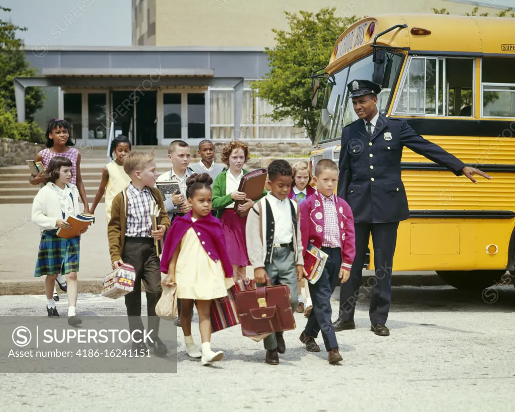 1960s GROUP OF ETHNICALLY DIVERSE ELEMENTARY STUDENTS AFTER SCHOOL CROSSING THE STREET PROTECTED BY AFRICAN-AMERICAN POLICEMAN