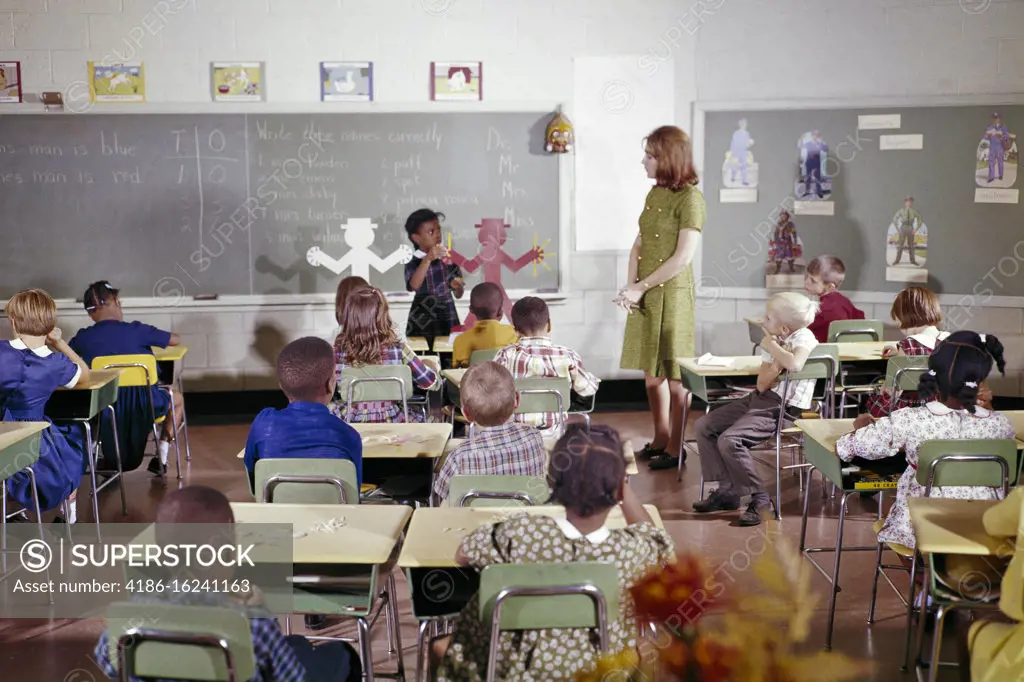1960s ELEMENTARY SCHOOL CLASSROOM CAUCASIAN TEACHER AND AFRICAN-AMERICAN STUDENT IN FRONT OF DIVERSE CLASS PRESENTING A LESSON