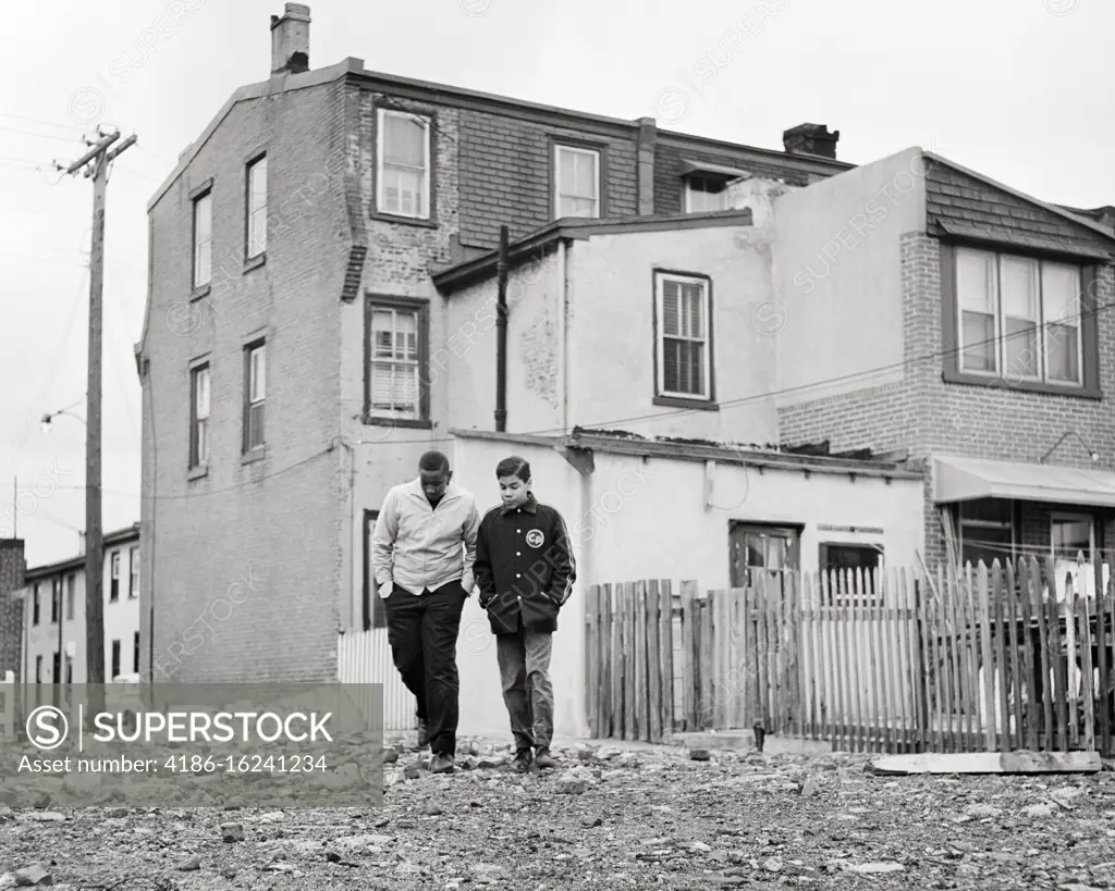 1960s TWO AT ODDS AFRICAN AMERICAN TEENAGE BOYS WALKING THROUGH BLIGHTED POVERTY STRICKEN URBAN AREA NEIGHBORHOOD 