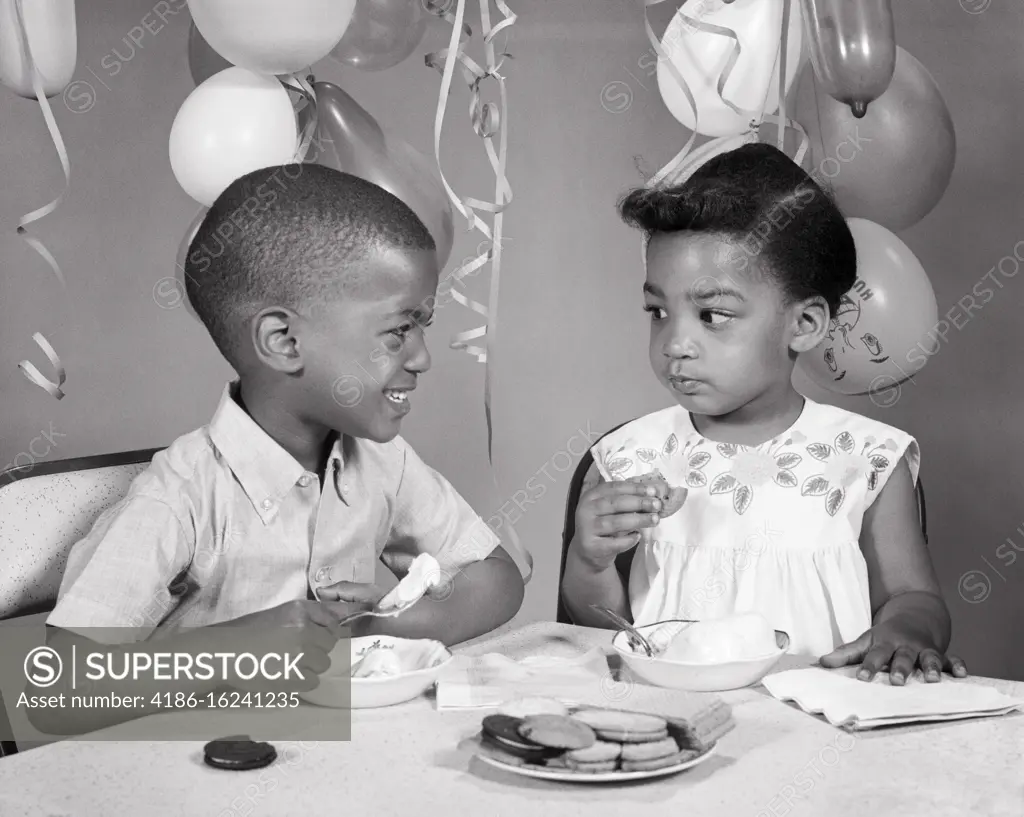 1960s TWO AFRICAN AMERICAN CHILDREN BOY GIRL BROTHER SISTER LOOKING AT EACH OTHER EATING ICE CREAM COOKIES BIRTHDAY PARTY