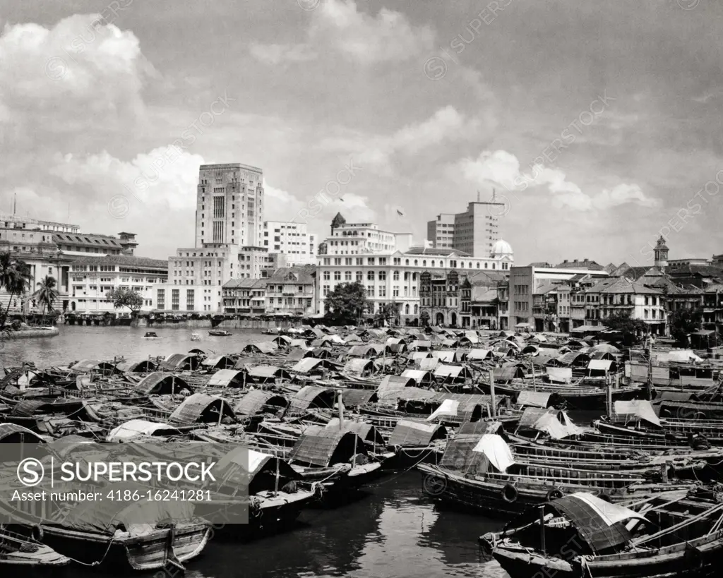 1950s 1960s CITY BUILDINGS SKYLINE SEEN ACROSS BUMBOAT CARGO BARGES MOORED IN HARBOR SINGAPORE