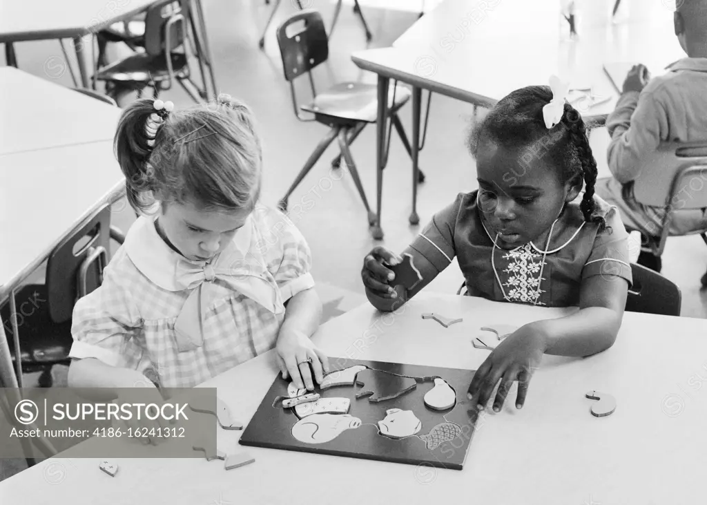 1960s TWO KINDERGARTEN PRESCHOOL GIRLS WORKING ON SOLVING A PUZZLE CAUCASIAN AND AFRICAN-AMERICAN