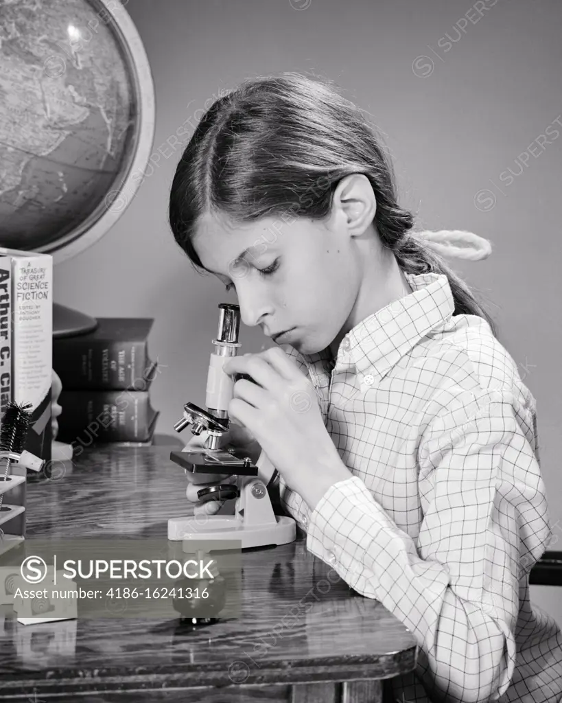 1960s ENGROSSED CURIOUS YOUNG SCHOOL GIRL AT HOME DESK USING A BEGINNER MICROSCOPE SURROUNDED BY GLOBE AND SCIENCE FICTION BOOKS