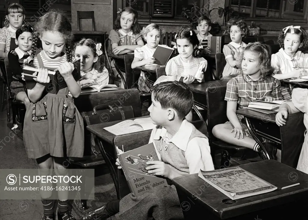 1950s ELEMENTARY SCHOOL CLASSROOM GIRL STUDENT STANDING BY DESK READING ALOUD FROM TEXTBOOK TO REST OF THE CLASS