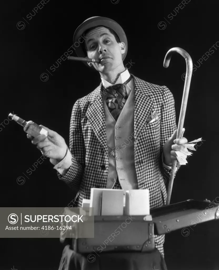 Carnival Barker Checkered Suit Cigar In Mouth Holding Cane Money In One Hand Bottle Of Tonic In Other Open Suitcase Stand 1930S