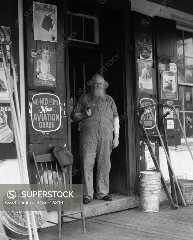 1930S Elderly Bearded Man In Overalls Smoking Pipe Standing In Doorway Of General Store