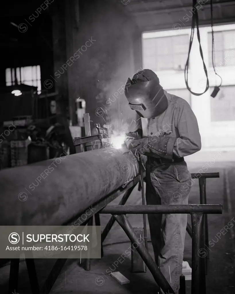 1960s ANONYMOUS MAN CONSTRUCTION WORKER ARC WELDING STEEL PIPE WEARING EYE AND VISION PROTECTING FACE MASK HELMET GLOVES