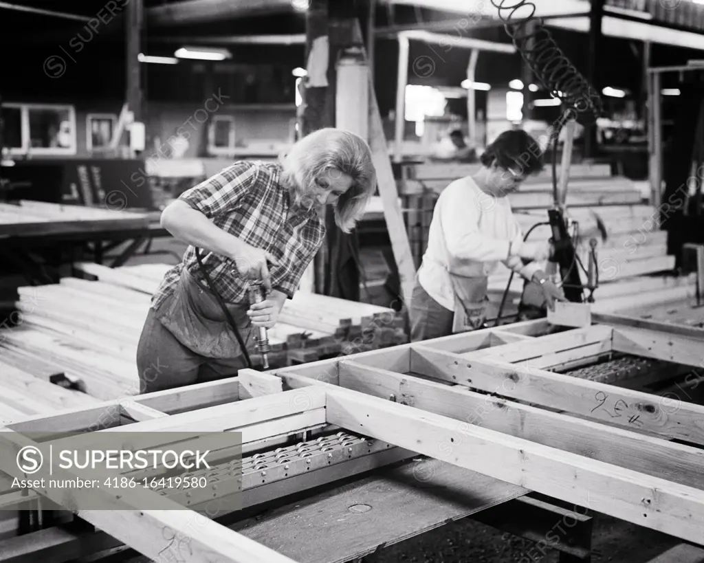 1960s 1970s TWO WORKERS WOMEN ON PREFAB FACTORY FLOOR ASSEMBLING A WOODEN FRAME FOR USE IN PREFABRICATED BUILDING 