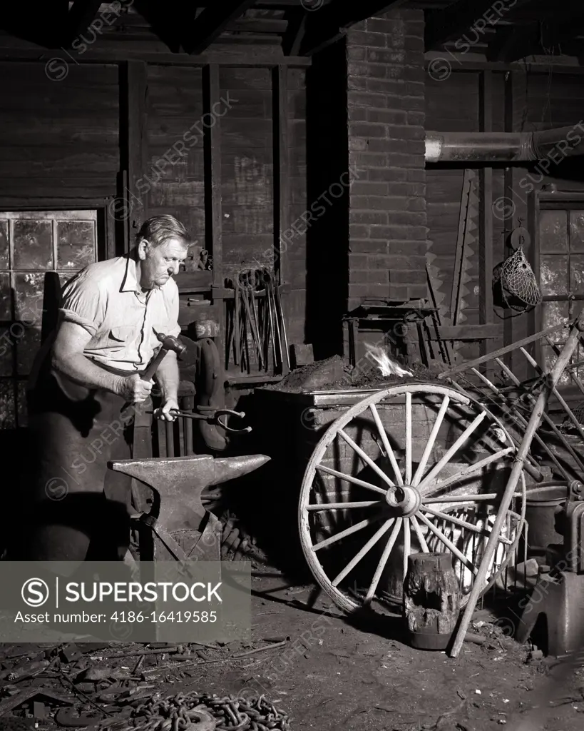 1950s 1960s MAN LOCAL VILLAGE BLACKSMITH WORKING AS FARRIER SHAPING A HORSESHOE WITH HAMMER ON ANVIL IN FORGE VIRGINIA USA