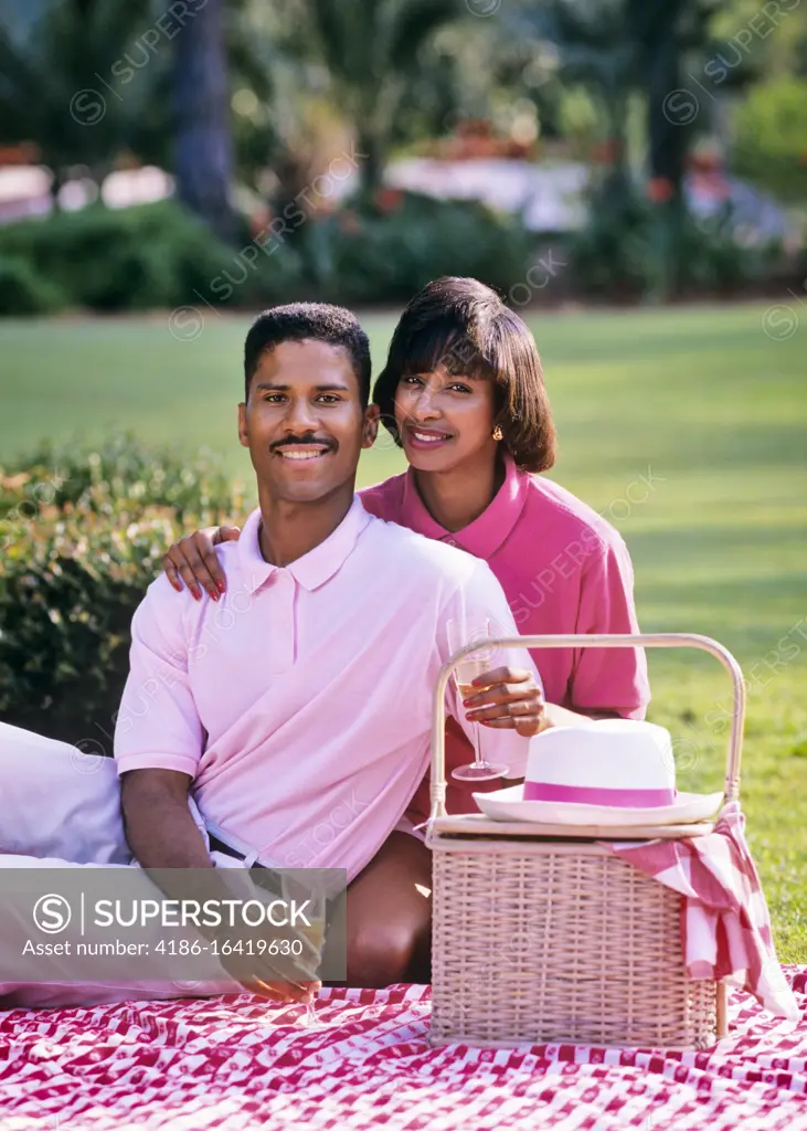 1980s 1990s AFRICAN-AMERICAN COUPLE SITTING LOOKING AT CAMERA ON BLANKET WITH PICNIC BASKET 