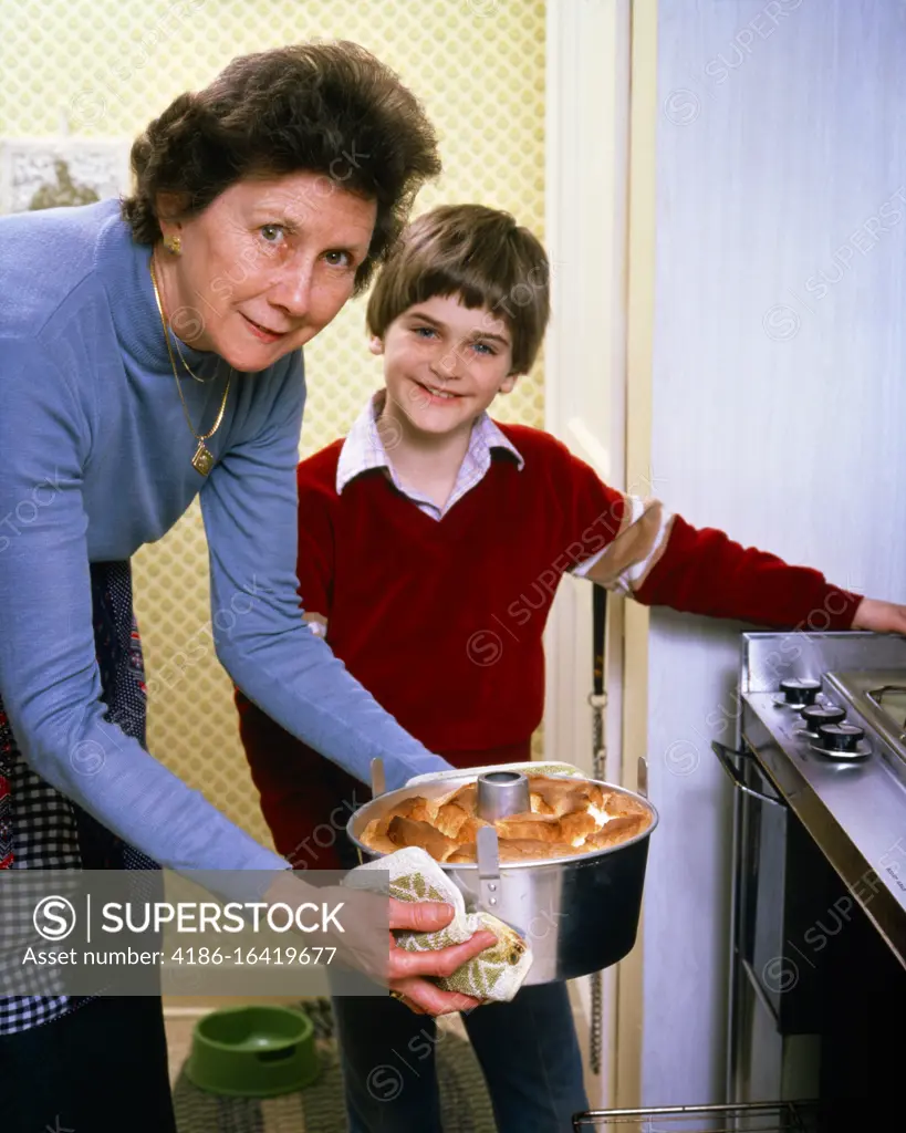 1980s GRANDMOTHER PROUDLY TAKING ANGEL FOOD CAKE OUT OF THE OVEN WITH GRANDSON STANDING BY BOTH SMILING BOTH LOOKING AT CAMERA