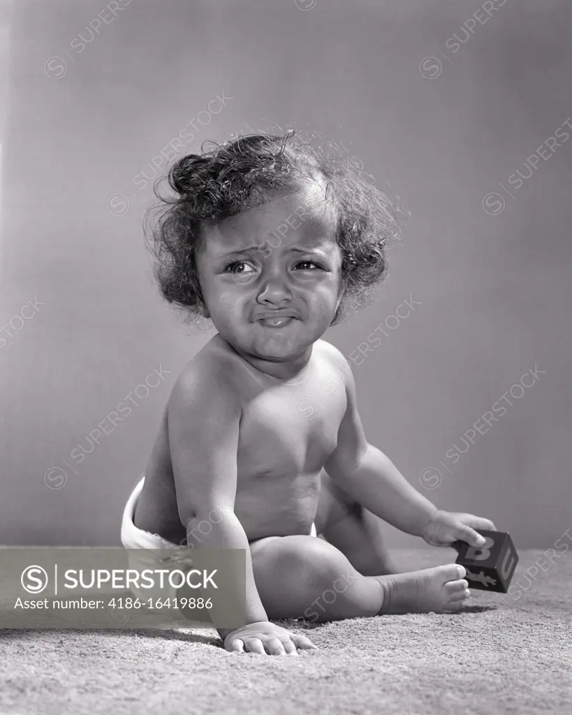 1940s 1950s UNHAPPY 1 YEAR OLD AFRICAN-AMERICAN BABY GIRL WEARING DIAPER SITTING PLAYING WITH TOY BLOCK GRIMACING LOOKING AWAY