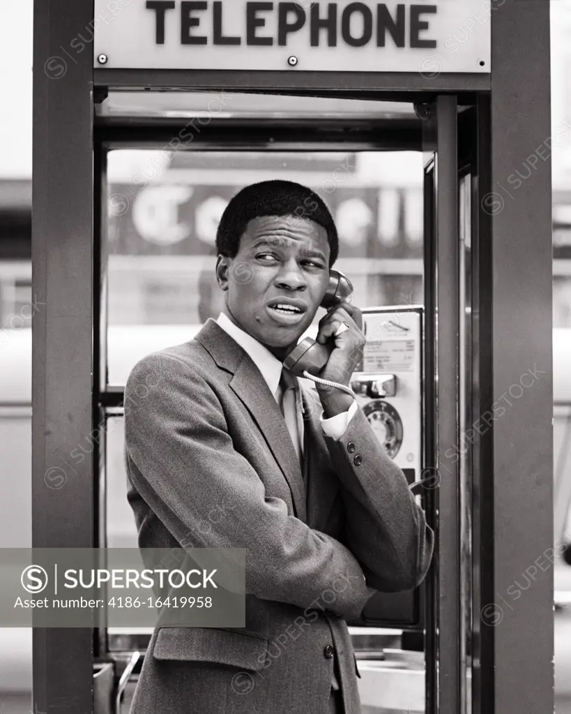 1970s ANXIOUS AFRICAN-AMERICAN MAN BUSINESSMAN AN UNSETTLED FACIAL EXPRESSION STANDING IN URBAN TELEPHONE BOOTH USING PAY PHONE