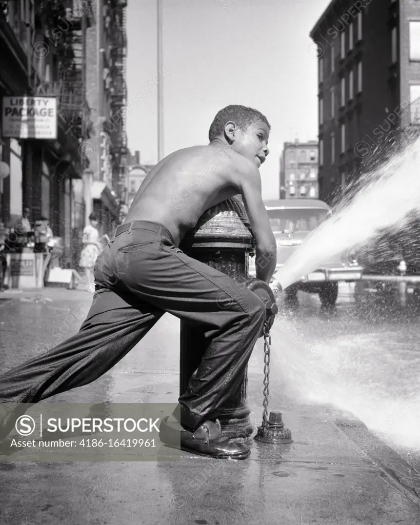 1970s ANONYMOUS TEENAGE AFRICAN-AMERICAN YOUNG MAN OPENING FIRE HYDRANT AND CREATING COOLING WATER SPRAY NEW YORK CITY NY USA