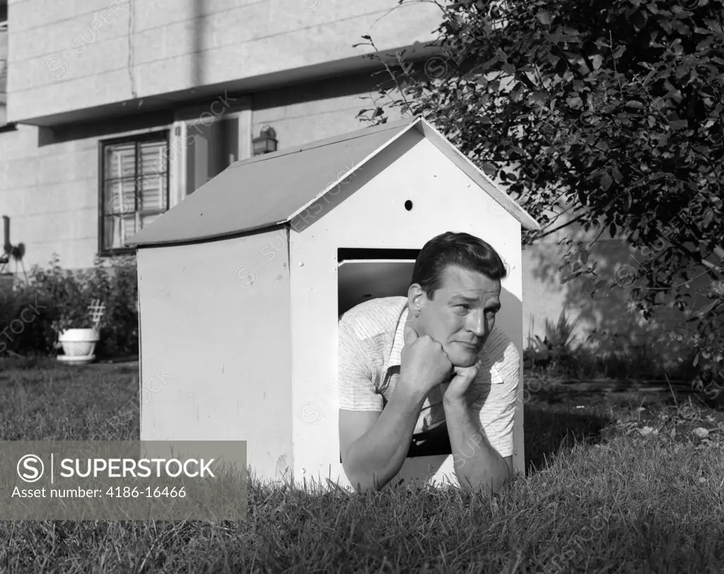 1960S Man In Doghouse In Backyard Resting On Elbows With Frown On Face