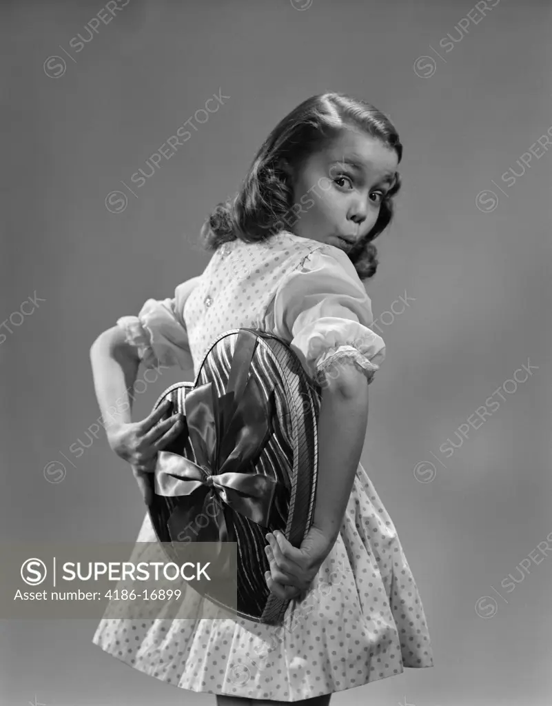 1950S Young Girl Holding Heart Shaped Candy Box Behind Back With Surprised Expression Wearing Polka Dot Dress