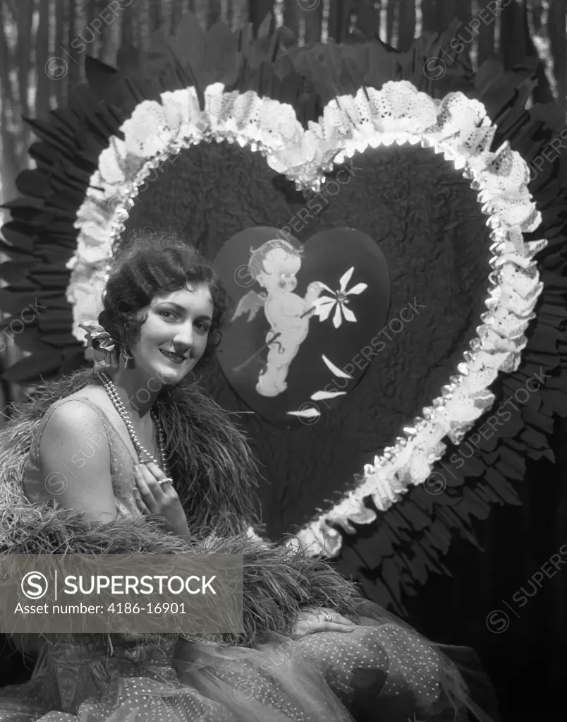 1920S 1930S Smiling Young Woman With Finger Wave Hair Pearls Feather Boa Party Dress Portrait In Front Of Giant Valentine Heart