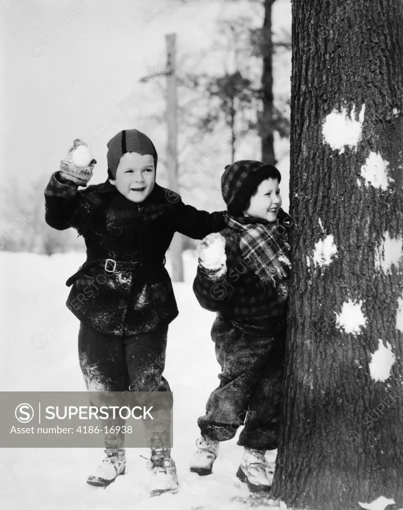 1920S 1930S Two Smiling Boys Playing In The Snow Hiding Behind Tree With Snowballs In Hand Have A Snowball Fight