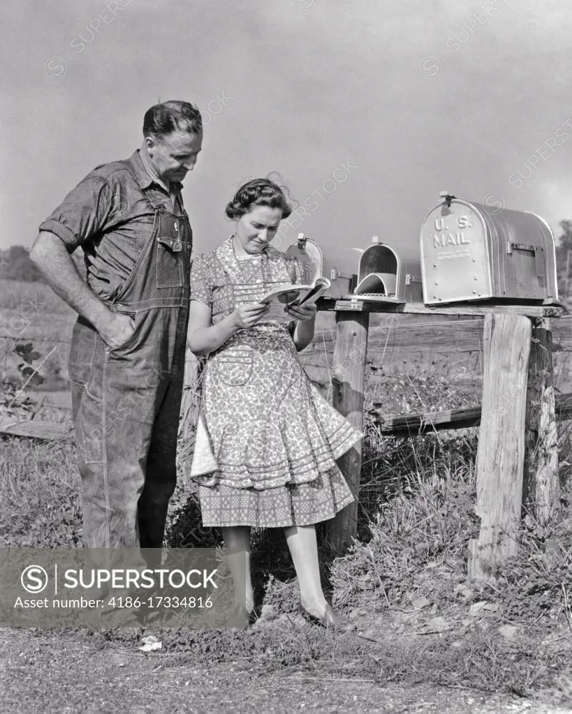 1930s 1940s MAN AND WOMAN FARMER AND WIFE TOGETHER COLLECTING MAIL FROM RURAL POSTAL MAILBOX READING MAGAZINE SEED CATALOG