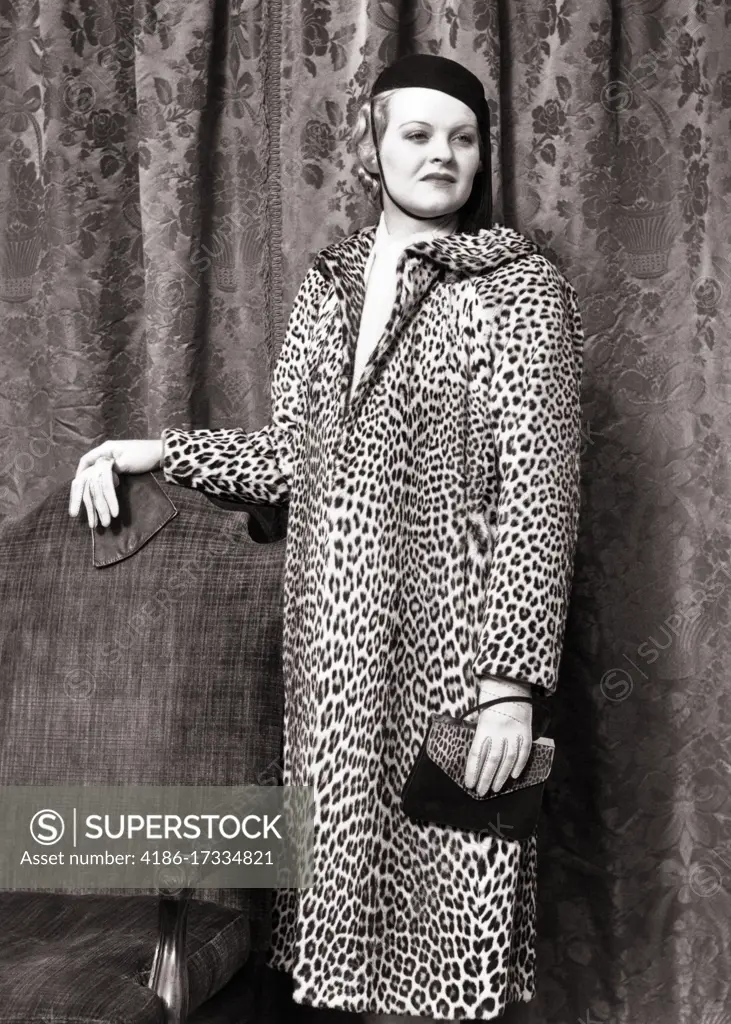 1930s 1938 FASHIONABLE WOMAN WEARING LEOPARD SKIN COAT STANDING BY CHAIR HOLDING ALLIGATOR HAND BAG WEARING LEATHER GLOVES