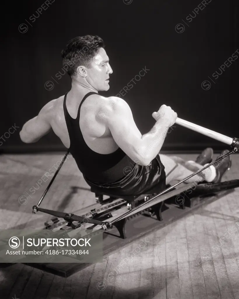 1930s MAN MUSCULAR ATHLETE CONCENTRATED FOCUSED FACIAL EXPRESSION EXERCISING WORKING OUT ON GYMNASIUM STATIONARY ROWING MACHINE