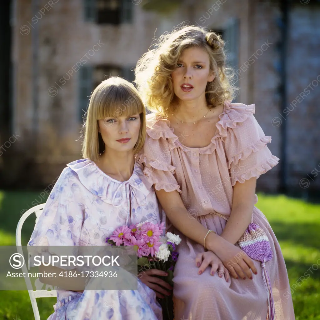 1970s 1980s TWO BLONDE WOMEN WEARING RUFFLED DRESSES IN PASTEL COLORS SITTING OUTDOORS LOOKING AT CAMERA 