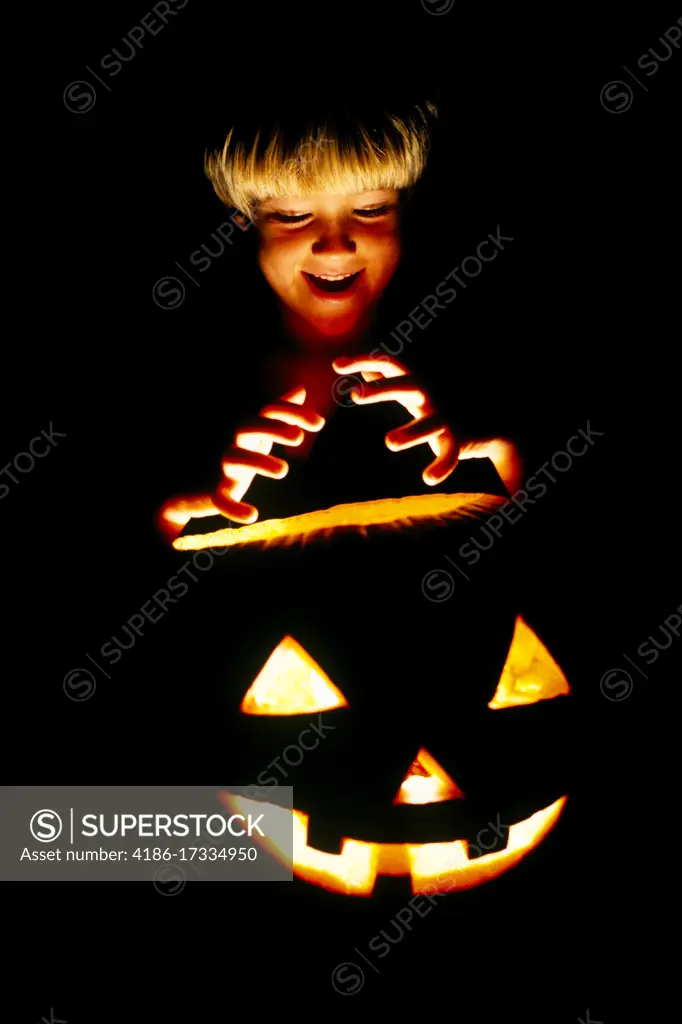 1980s BLONDE BOY IN SCARY POSE MAKING A THREATENING HAND GESTURE ABOVE AN ILLUMINATED HALLOWEEN CARVED PUMPKIN AT NIGHT