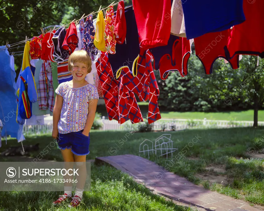 1970s LITTLE GIRL BAND-AID ON KNEE SMILING LOOKING AT CAMERA STANDING IN BACKYARD BEFORE CLOTHESLINE WITH VERY COLORFUL LAUNDRY