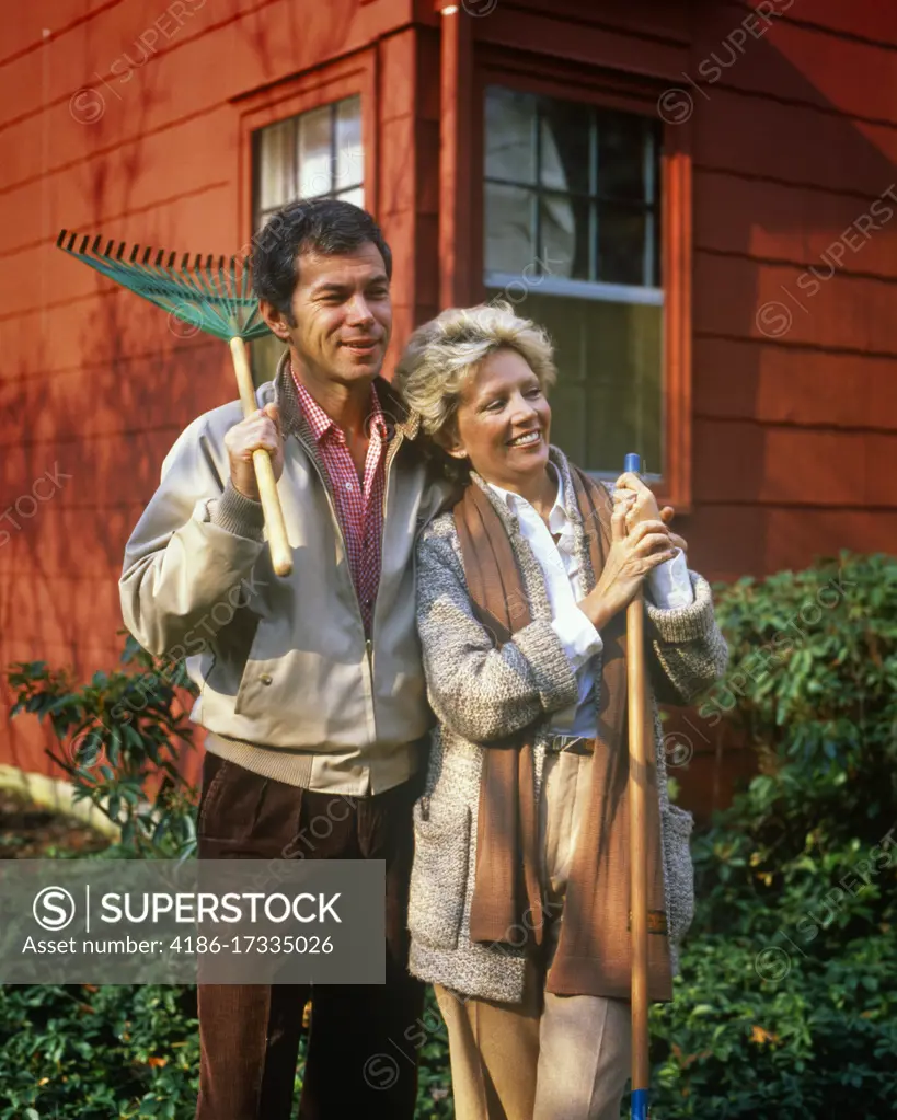 1980s MATURE COUPLE STANDING BY HOUSE MAN HOLDING RAKE ON SHOULDER WOMAN LEANING ON GARDEN TOOL WEARING AUTUMN CLOTHES SMILING