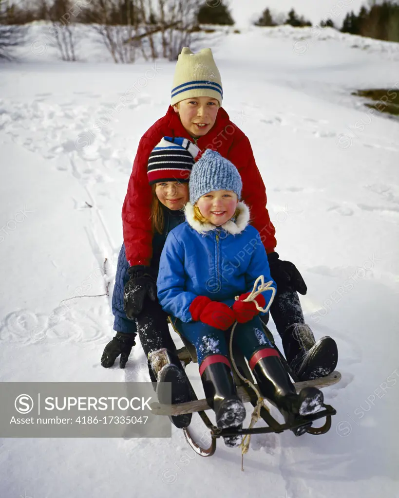 1960s PRETEEN BOY RIDING ON A SNOW SLED WITH HIS TWO YOUNGER SISTERS SMILING LOOKING AT CAMERA ALL WEARING KNIT WOOL HATS